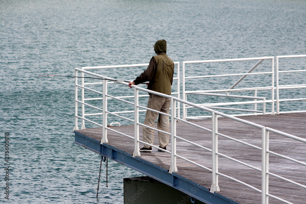 Man in hunting jacket and hoodie standing at the end of local pier fishing with small fishing rod over white metal fence on rainy spring day