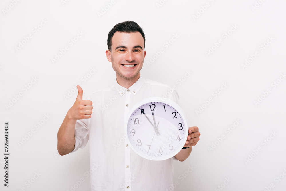 Joyful young man is holding a clock and showing thumb up in studio on white background. Joyful young man is holding a clock and showing thumb up in studio on white background.
