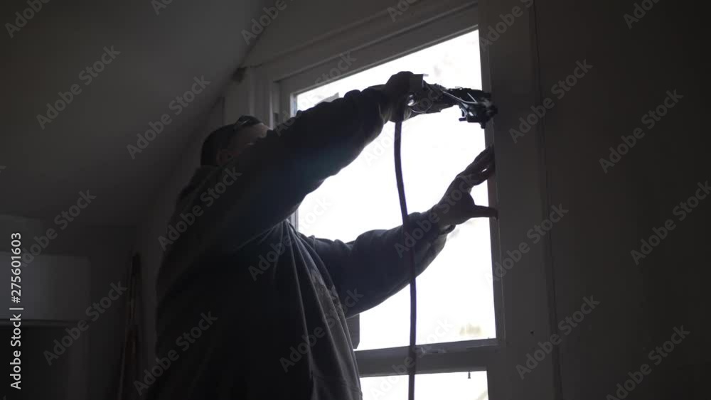 A man works inside remodeling a house - uses nail gun to hang window trim. A man works inside remodeling a house - uses nail gun to hang window trim.