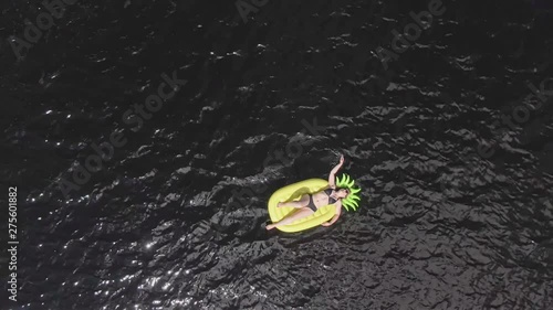 Top aerial pull out with rotation of blonde Swedish woman relaxing on an inflatable pineapple floating on a dark lake in Nacka, Stockholm. photo
