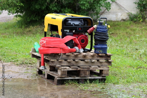 New large petrol water pump next to small blue electrical water pump and thick red hose all put on wooden pallets waiting to be used for pumping flood water away from family houses backyards on rainy 