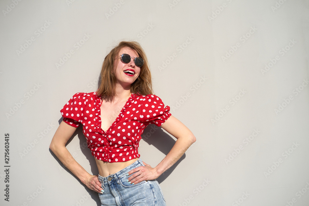 Redhead caucasian girl in red polka-dot pattern blouse and jeans smiles staying near grey background Redhead caucasian girl in red polka-dot pattern blouse and jeans smiles staying near grey background