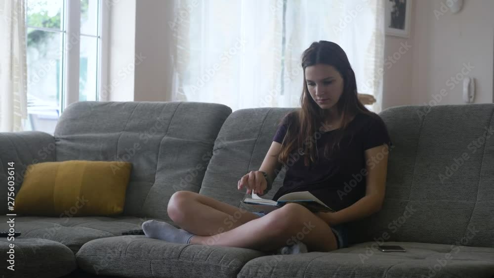 Static wide shot of a lady sitting on a sofa reading a book then replacing it with her cellphone and smiles Static wide shot of a lady sitting on a sofa reading a book then replacing it with her cellphone and smiles