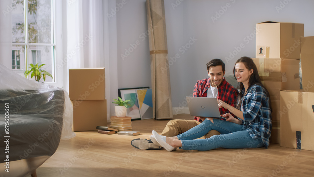 Happy Young Couple Sitting on the Floor of the Newly Rented / Purchased Apartment Use Laptop Computer. Unpacked Cardboard Boxes and Covered Furniture in the Modern Bright Sweet Home. Happy Young Couple Sitting on the Floor of the Newly Rented / Purchased Apartment Use Laptop Computer. Unpacked Cardboard Boxes and Covered Furniture in the Modern Bright Sweet Home.