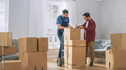 Happy New Homeowner Welcomes Professional Mover with Hand Truck full of Cardboard Boxes, Receives His Goods and Signs on Clipboard. photo