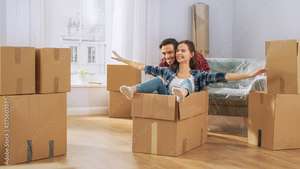 Happy Young Couple Moving Into New Apartment, Boyfriend Drives Girlfriend in Cardboard Box, She imitates Airplane Wings. Young Boyfriend, Girlfriend Start Living Together, Unpacking Stuff, Having Fun. Happy Young Couple Moving Into New Apartment, Boyfriend Drives Girlfriend in Cardboard Box, She imitates Airplane Wings. Young Boyfriend, Girlfriend Start Living Together, Unpacking Stuff, Having Fun.
