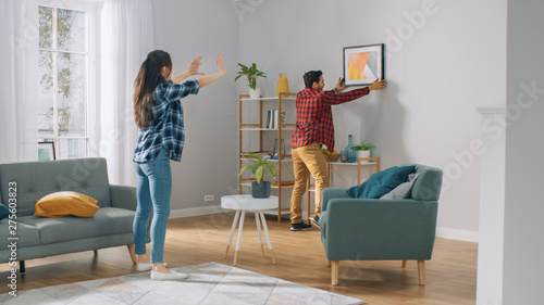 Obraz na płótnie Happy Couple Hanging Picture on the Wall, Boyfriend Moves It, Girlfriend Tells Him when the Frame is Hanging Straight