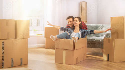 Young Couple Moving Into New Apartment, Boyfriend Drives Girlfriend in Cardboard Box She imitates Airplane Wings. Young Boyfriend and Girlfriend Start Living Together. Room Lit by Warm Yellow Sunlight