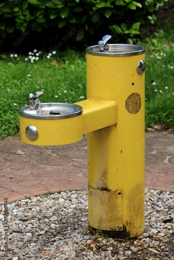 Public park old dilapidated yellow two level drinking fountain for adults and children surrounded with gravel and stone tiles on warm sunny spring day Public park old dilapidated yellow two level drinking fountain for adults and children surrounded with gravel and stone tiles on warm sunny spring day