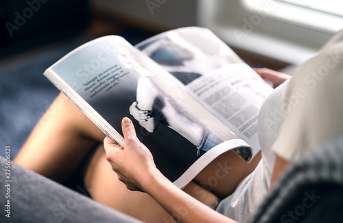 Happy millennial lady reading fashion magazine with latest beauty trends or celebrity news and interview articles. Woman sitting on couch with open page. Young person relaxing and enjoying fun leisure photo