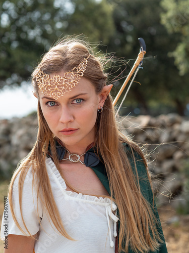 Fotografía Blonde girl with elf ears poses in the field with a bow and a green cape