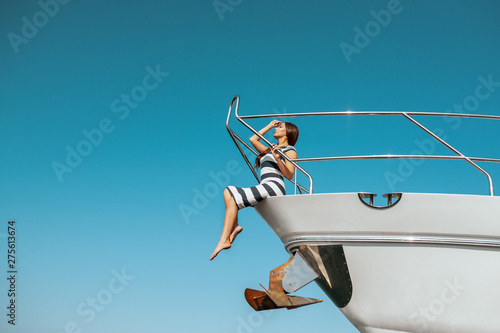 Fashion photo of adorable young woman in striped dress sitting on edge of luxury yacht and looking for the sea during sailing trip. Happy woman enjoying summer travel. Vacation or holiday concept © Detkov D