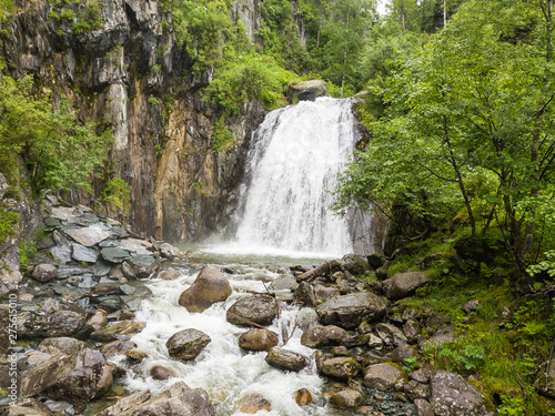A large waterfall in the back of the Altai Mountains with gray-brown stones near a steep cliff with green trees. Rest and loneliness while traveling to deserted places.