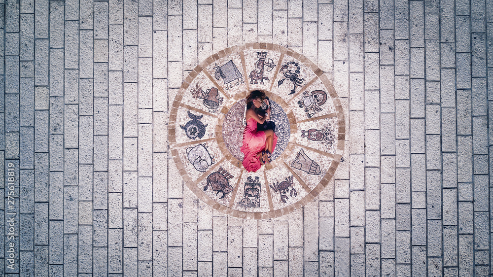 Aerial photo: A charming lady in a pink dress is lying on a stone pavement, zodiac signs are depicted. Horoscope.