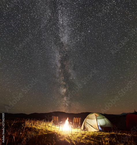 Panoramic view of tourist camping at night in the mountains. Glowing tent, two chairs and bonfire under amazing night sky full of stars and Milky way. Tourism active lifestyle astrophotography concept