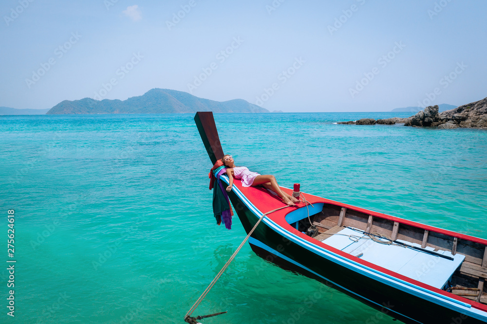 Aerial drone shot of a young blonde woman in a pink dress and sunglasses in the front of a wooden longtail thai boat. Crystal clear water and corals at a tropical island and amazing beach. Aerial drone shot of a young blonde woman in a pink dress and sunglasses in the front of a wooden longtail thai boat. Crystal clear water and corals at a tropical island and amazing beach.