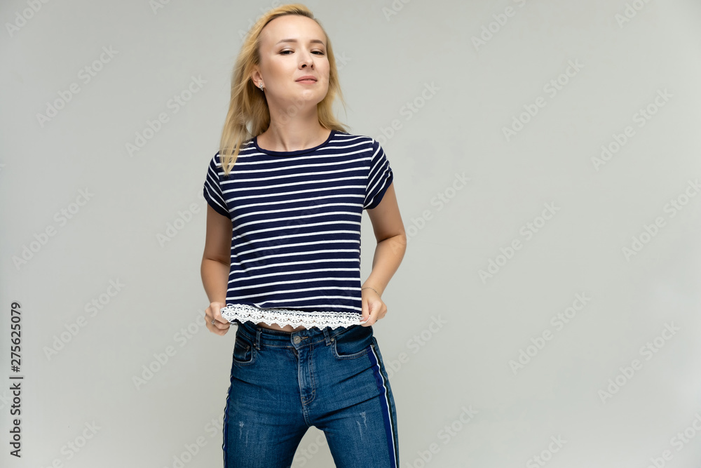 Portrait of a pretty interesting blonde girl on a white background in jeans and a striped blue t-shirt. Standing in front of the camera, smiling, showing hands. Shows a lot of emotions. Portrait of a pretty interesting blonde girl on a white background in jeans and a striped blue t-shirt. Standing in front of the camera, smiling, showing hands. Shows a lot of emotions.