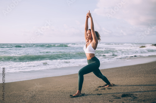 Professional fitness girl doing yoga exercise outdoors to maintain vital tone and develop flexibility, young woman dressed in athletic apparel standing in special warrior pose during morning pilates