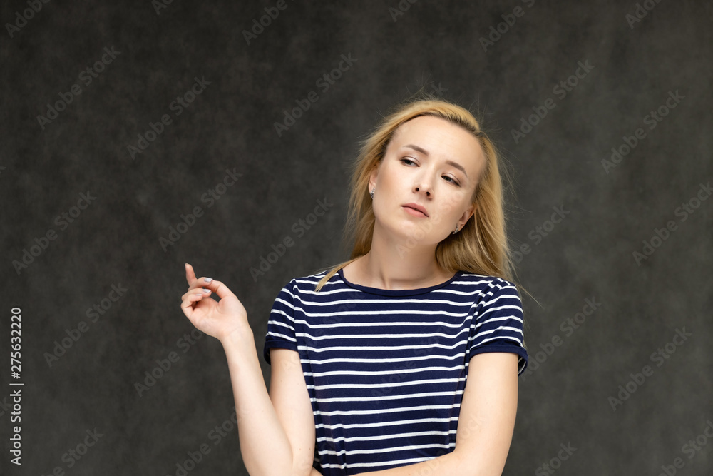 Portrait to the waist of a pretty interesting fashionable blonde girl on a gray background in a striped blue t-shirt. Standing in front of the camera, smiling, showing hands. Shows a lot of emotions. Portrait to the waist of a pretty interesting fashionable blonde girl on a gray background in a striped blue t-shirt. Standing in front of the camera, smiling, showing hands. Shows a lot of emotions.
