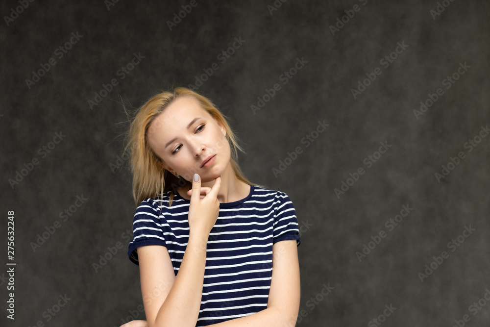 Portrait to the waist of a pretty interesting fashionable blonde girl on a gray background in a striped blue t-shirt. Standing in front of the camera, smiling, showing hands. Shows a lot of emotions. Portrait to the waist of a pretty interesting fashionable blonde girl on a gray background in a striped blue t-shirt. Standing in front of the camera, smiling, showing hands. Shows a lot of emotions.