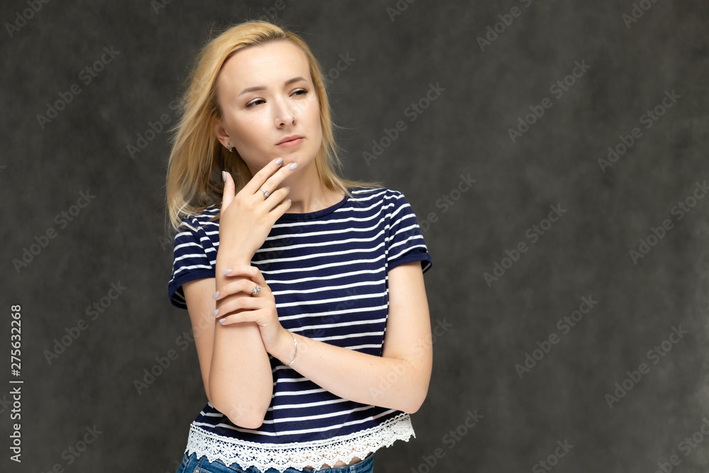 Portrait to the waist of a pretty interesting fashionable blonde girl on a gray background in a striped blue t-shirt. Standing in front of the camera, smiling, showing hands. Shows a lot of emotions. Portrait to the waist of a pretty interesting fashionable blonde girl on a gray background in a striped blue t-shirt. Standing in front of the camera, smiling, showing hands. Shows a lot of emotions.