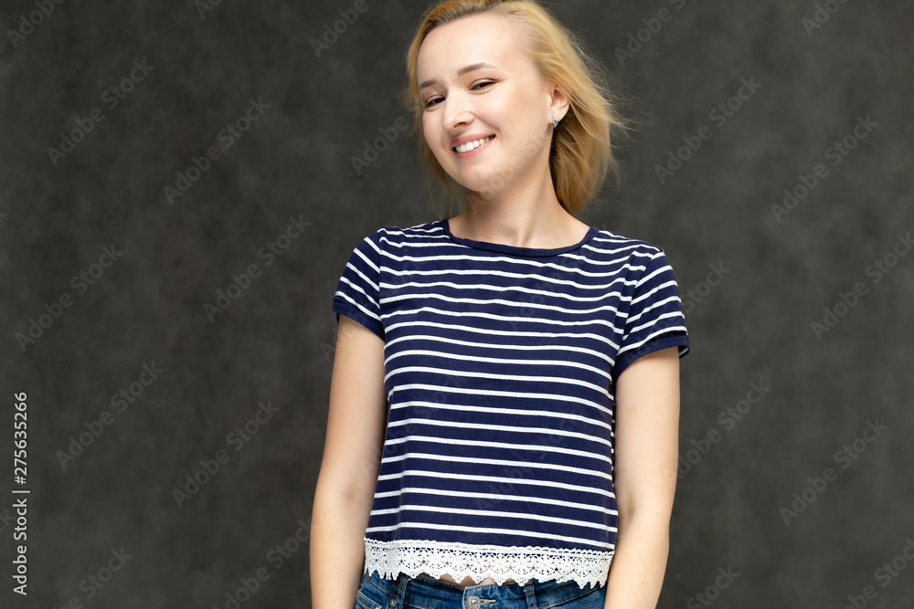 Portrait to the waist of a pretty interesting fashionable blonde girl on a gray background in a striped blue t-shirt. Standing in front of the camera, smiling, showing hands. Shows a lot of emotions. Portrait to the waist of a pretty interesting fashionable blonde girl on a gray background in a striped blue t-shirt. Standing in front of the camera, smiling, showing hands. Shows a lot of emotions.
