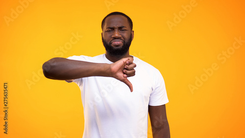 Disappointed afro-american guy showing thumbs down on yellow background, hater