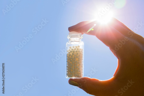 Selective focus on person hand holding glass jar full of small white round homeopathy pills against blue sky background. photo