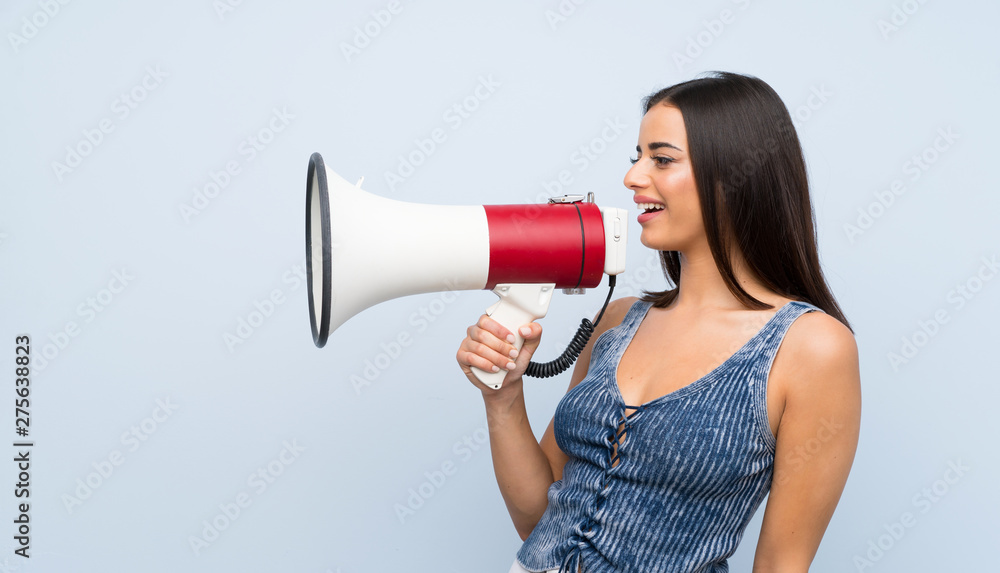 Young woman over isolated blue wall shouting through a megaphone Young woman over isolated blue wall shouting through a megaphone