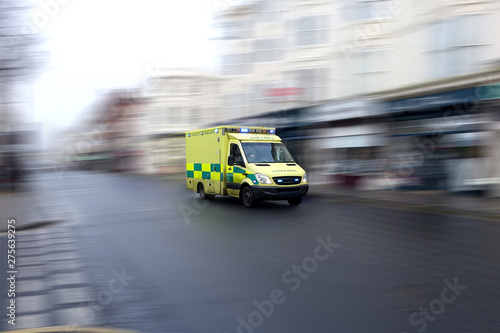 Yellow emergency ambulance rushing through a city to the hospital with full flashing blue lights and siren. Speed is emphasized by the motion blurred urban background. Vehicle is generic: no visible m