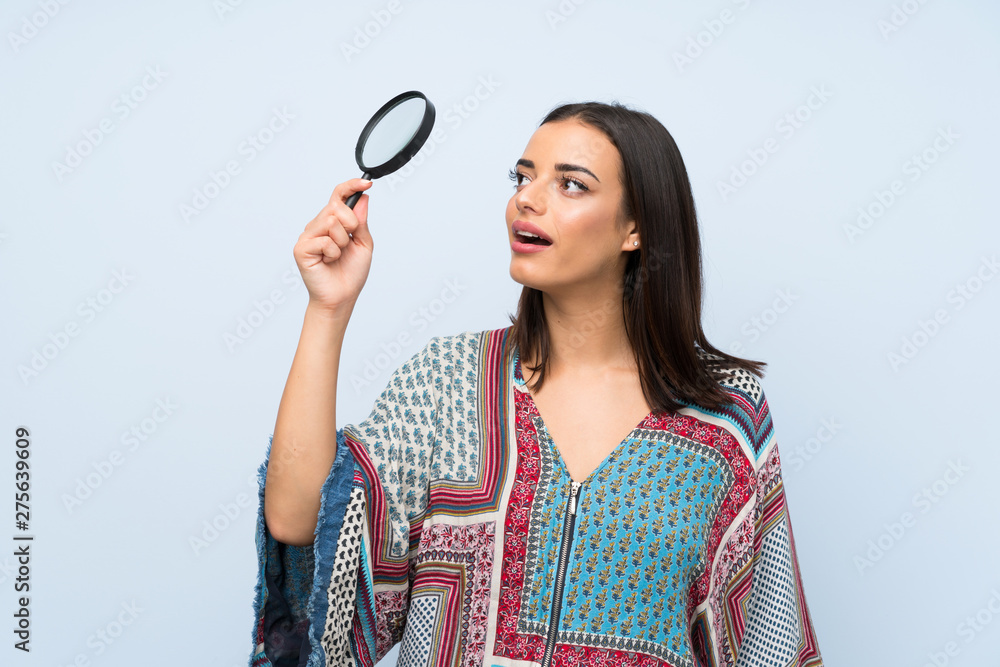 Young woman over isolated blue wall holding a magnifying glass Young woman over isolated blue wall holding a magnifying glass