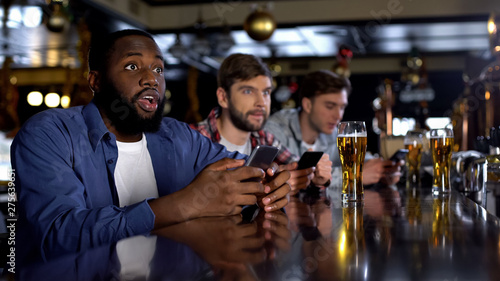 African-american man checking bets on smartphone, watching match in bar, app