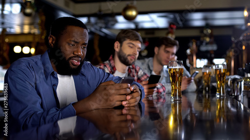 Sad multiethnic men surfing internet on phones in pub instead of communicating