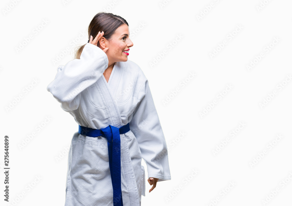 Young beautiful woman wearing karate kimono uniform over isolated background smiling with hand over ear listening an hearing to rumor or gossip. Deafness concept. Young beautiful woman wearing karate kimono uniform over isolated background smiling with hand over ear listening an hearing to rumor or gossip. Deafness concept.