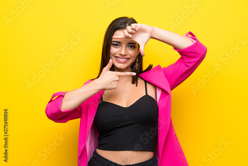 Young woman over isolated yellow background focusing face. Framing symbol