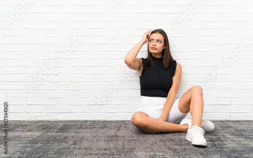 Pretty young woman sitting on the floor having doubts while scratching head