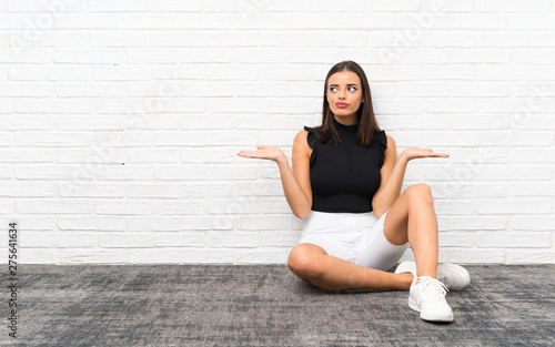 Pretty young woman sitting on the floor having doubts while raising hands