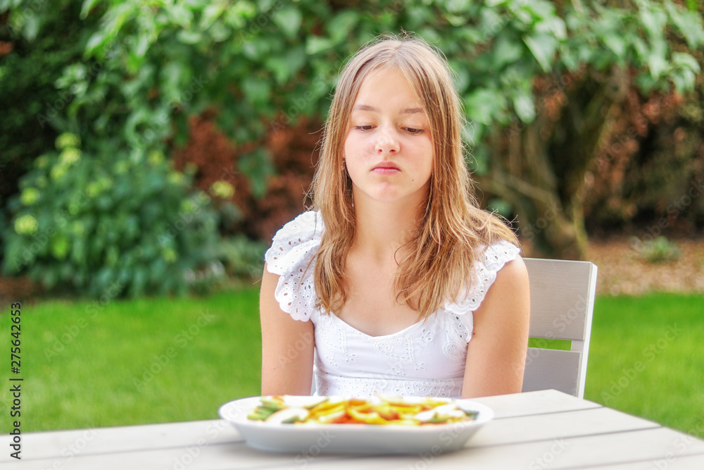 Pretty preteen looking at dried fruit and vegetable chips with unhaddy face sitting in garden outdoors at summer day. Doesn’t like healthy snack. Summer lifestyle