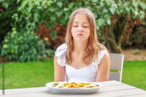 Pretty preteen looking at dried fruit and vegetable chips with unhaddy face sitting in garden outdoors at summer day. Doesn’t like healthy snack. Summer lifestyle