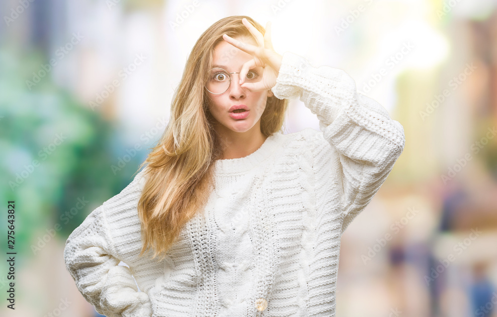 Young beautiful blonde woman wearing winter sweater and sunglasses over isolated background doing ok gesture shocked with surprised face, eye looking through fingers. Unbelieving expression.