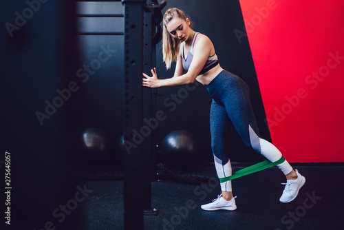 Crossfit and healthy lifestyle concept, charming beautiful sporty muscular fitness model lady dressed in leggins and top training with green elastic band on legs muscles doing cardio exercise