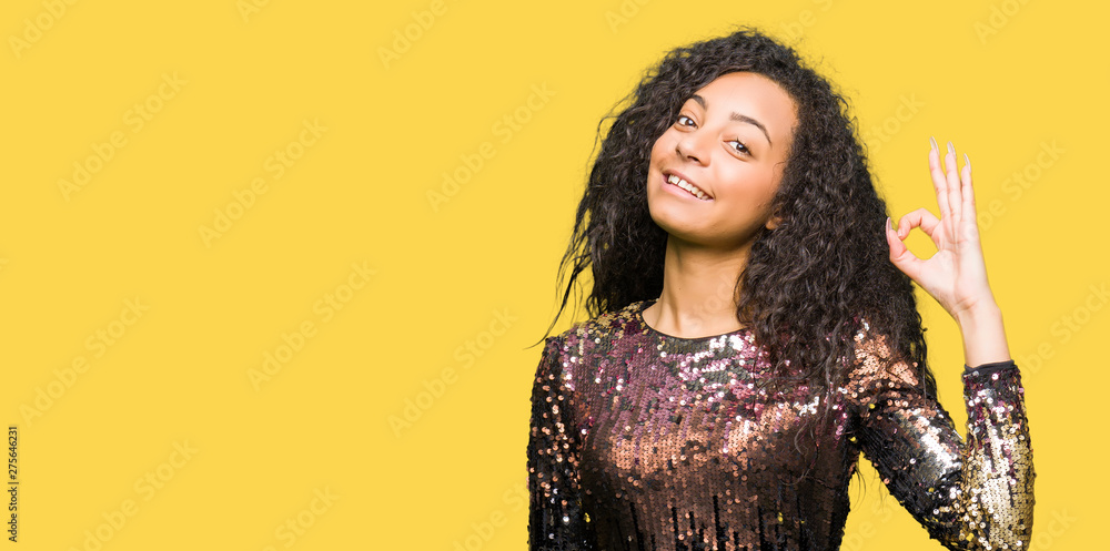 Young beautiful girl with curly hair wearing night party dress smiling positive doing ok sign with hand and fingers. Successful expression. Young beautiful girl with curly hair wearing night party dress smiling positive doing ok sign with hand and fingers. Successful expression.