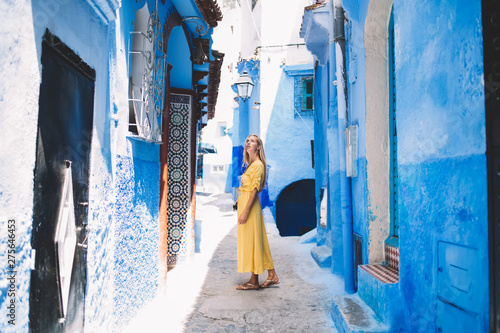 Attractive hipster girl standing in middle of ancient medina street - Morocco looking around discovering country, pensive woman in yellow dress thinking about traditional arabic town with blue walls © BullRun