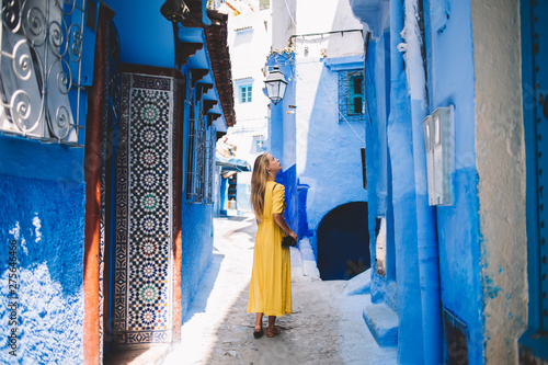 Beautiful girl with amateur camera standing in tourists city - Morocco inspecting oriental mosaic and blue walls, back view of Caucasian woman walking between ancient houses in old arabic medina © BullRun
