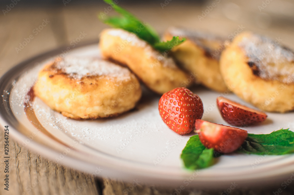 Gourmet breakfast - cottage cheese pancakes, cheesecakes, cottage cheese pancakes with strawberries, mint and powdered sugar in a white plate. Useful dessert on a wooden table in rustic style. Selecti
