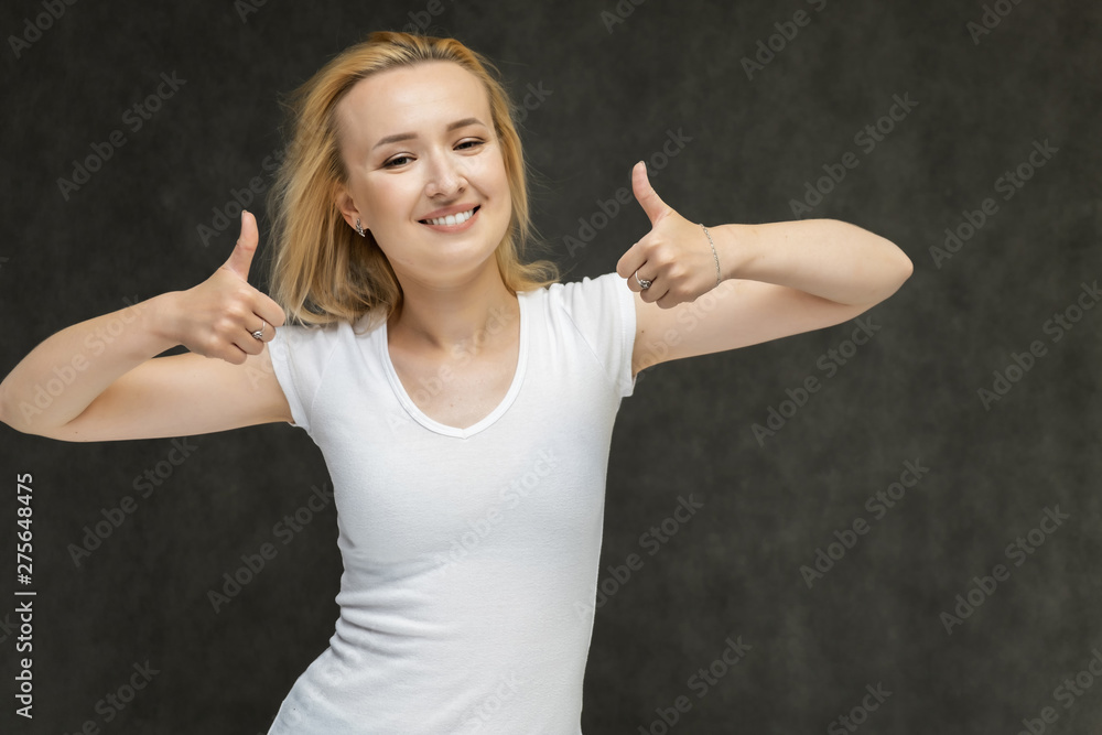 Waist up portrait of pretty interesting fashionable blonde girl on gray background in white t-shirt. Standing in front of the camera, smiling, showing hands. Shows a lot of emotions. Waist up portrait of pretty interesting fashionable blonde girl on gray background in white t-shirt. Standing in front of the camera, smiling, showing hands. Shows a lot of emotions.