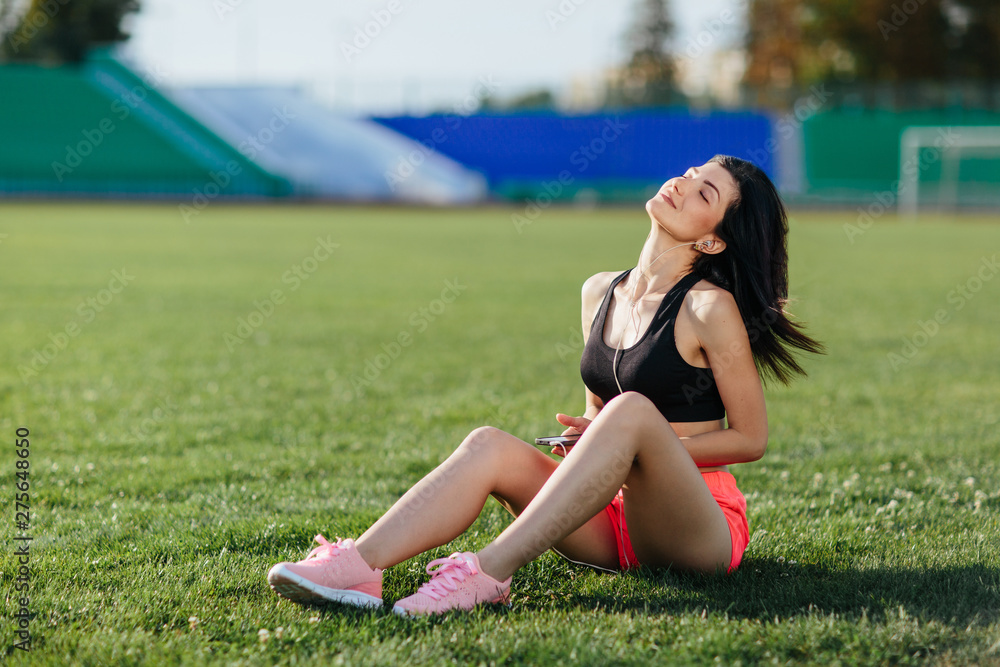 Young sporty woman brunette in sport dress sitting on the grass football field stadium and listens to music in earphones, her head thrown back, she gets pleasure, relaxes. Sport and healthy lifestyle