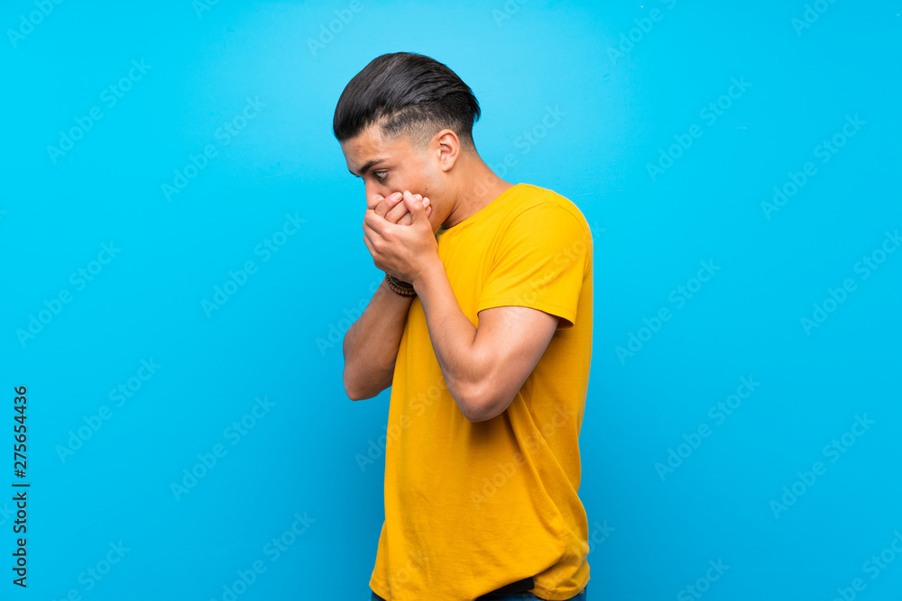 Young man with yellow shirt over isolated blue background covering mouth and looking to the side Young man with yellow shirt over isolated blue background covering mouth and looking to the side