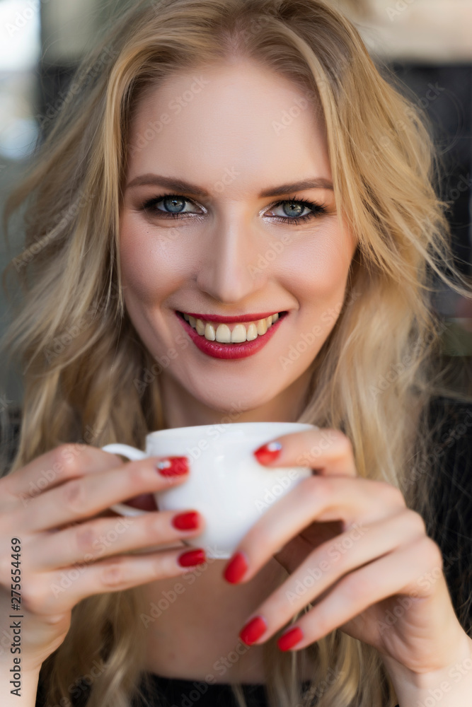 Beautiful smiling long haired blonde girl sits and rests at the table in cafe, holding a cup of coffee in her hands. Close up. Trendy, commercial design. Copy space. Beautiful smiling long haired blonde girl sits and rests at the table in cafe, holding a cup of coffee in her hands. Close up. Trendy, commercial design. Copy space.