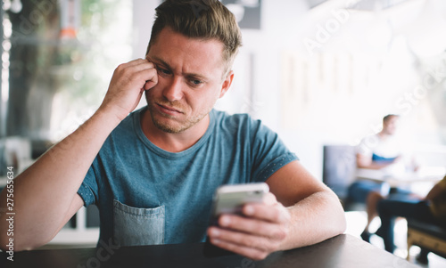 Puzzled male blogger feeling tired from online messaging with followers from own website using smartphone application during leisure in cafeteria, concept of millennial people and generation